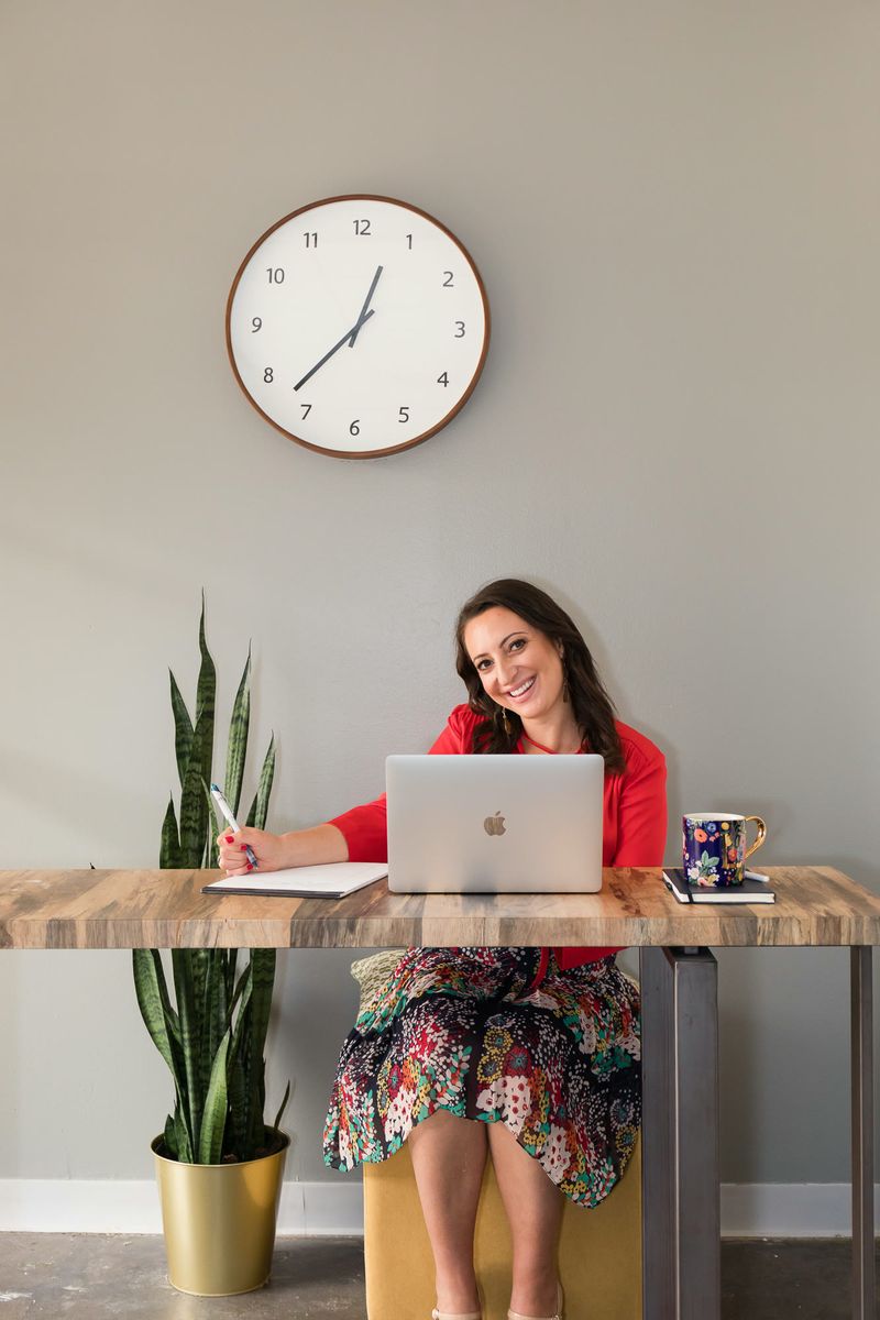 Lauren Goldstein working at her desk with a laptop and notebook, smiling, beneath a wall clock